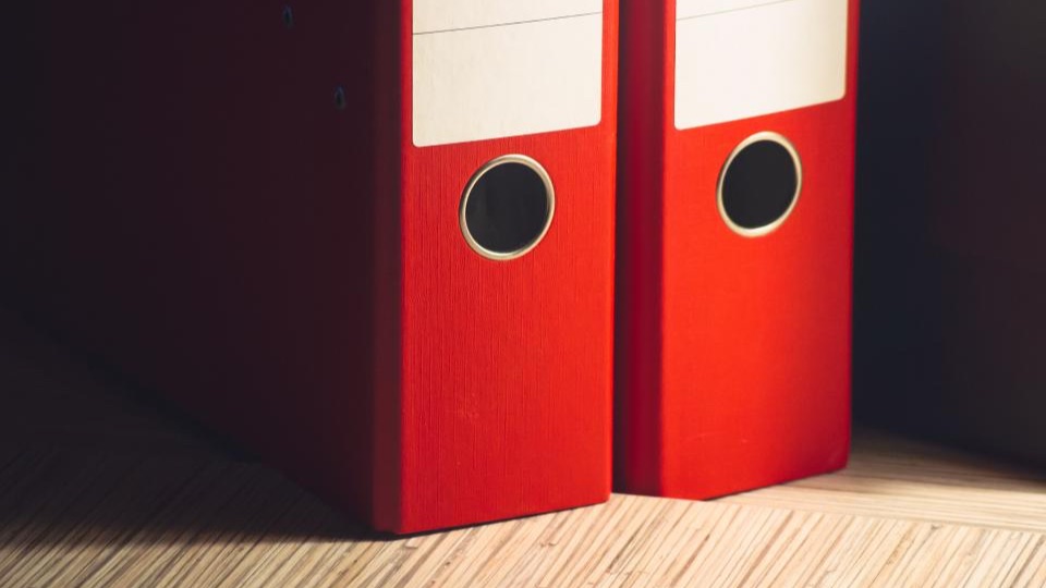 Two red lever arch files with blank labels standing on a light wooden floor