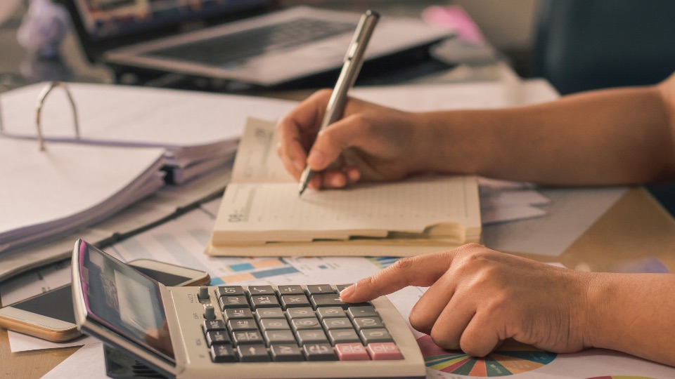 Hands writing in a notebook beside a calculator and printed reports on a desk