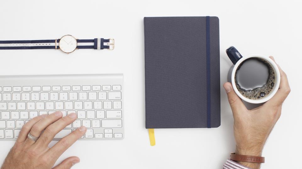 Overhead view of a closed navy notebook between a keyboard and a cup of coffee on a white desk